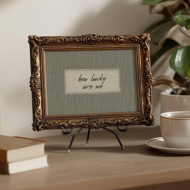 May include: Ornate gold-colored picture frame with a green and white checkered background. The frame holds a fabric rectangle with the text "how lucky are we". The frame is propped up on a black metal stand.