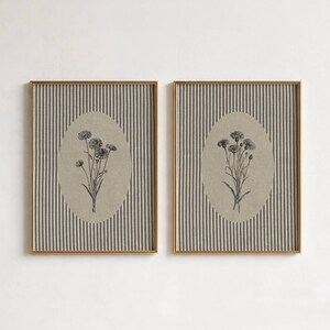 May include: Two rectangular wall art prints in thin gold frames. Each print features a vertical dark gray and white pinstripe background with a central beige oval containing a detailed black ink botanical illustration of a small flower bouquet.