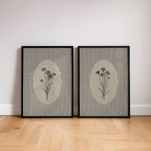 May include: Two rectangular framed wall art pieces featuring a black and white botanical sketch of wildflowers centered within an oval beige background, set against a backdrop of vertical grey and white pinstripes inside a black frame.