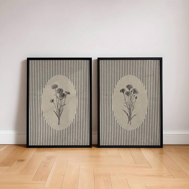 May include: Two rectangular framed wall art pieces featuring a black and white botanical sketch of wildflowers centered within an oval beige background, set against a backdrop of vertical grey and white pinstripes inside a black frame.