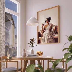 Puede incluir: Una obra de arte enmarcada representa a una mujer con un sari blanco sosteniendo una copa de vino. La obra de arte se exhibe en una pared blanca en un comedor. Una mesa redonda de madera y sillas est&aacute;n en primer plano, con una ventana y una l&aacute;mpara colgante.