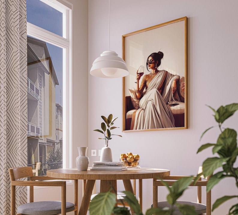 Puede incluir: Una obra de arte enmarcada representa a una mujer con un sari blanco sosteniendo una copa de vino. La obra de arte se exhibe en una pared blanca en un comedor. Una mesa redonda de madera y sillas est&aacute;n en primer plano, con una ventana y una l&aacute;mpara colgante.