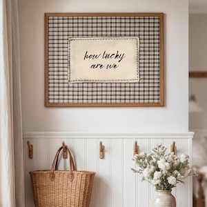 May include: A rectangular wall art piece featuring a brown and white plaid fabric background and a centered cream-colored fabric patch with the black text "how lucky are we" stitched onto the fabric, all enclosed in a light brown wooden frame.