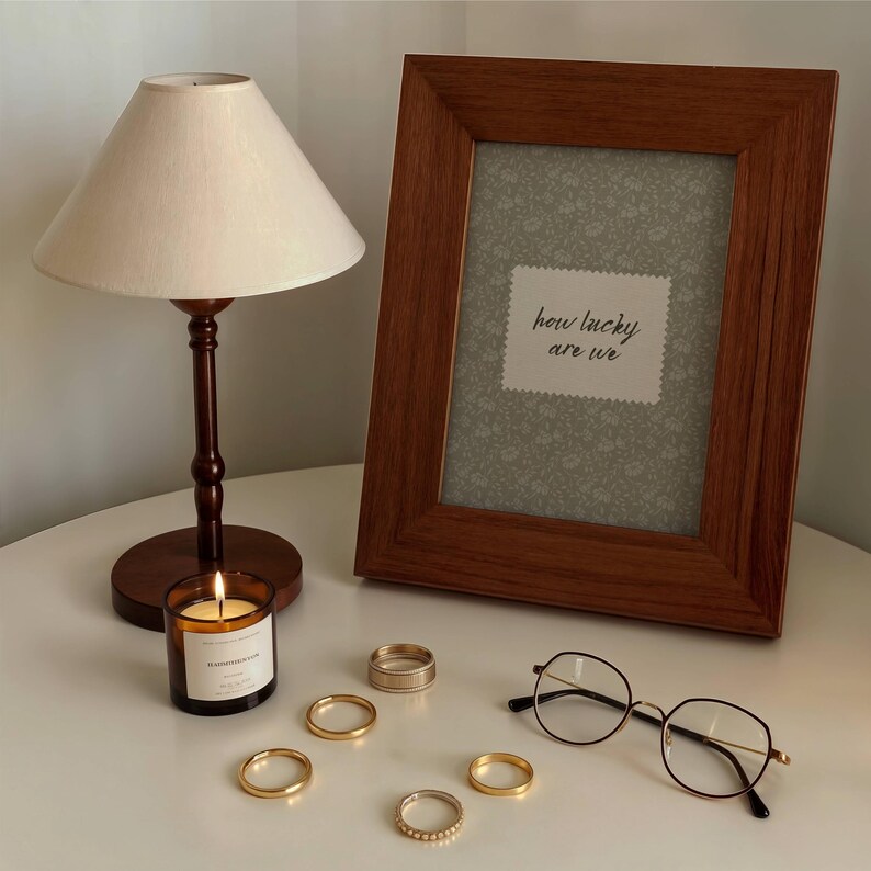 May include: A lit candle in a glass jar, a wooden framed picture with the text "how lucky are we", a small table lamp, several gold rings, and round eyeglasses with a gold frame are arranged on a white surface.