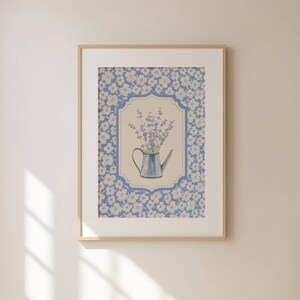 May include: Framed art print featuring a watercolor illustration of lavender in a blue watering can. The artwork is set against a floral patterned background in shades of blue and white, framed in a light wood.