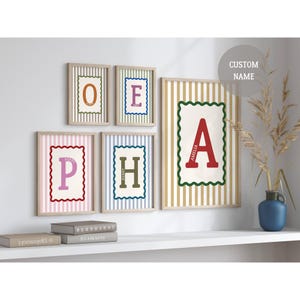 Puede incluir: Colección de cuadros con letras personalizadas. Cada cuadro presenta una letra en un color llamativo sobre un fondo crema con un borde decorativo. Los marcos están dispuestos en una estantería blanca, creando una decoración mural personalizada.