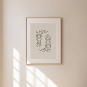 May include: A framed print showing a minimalist illustration of two cowboy boots. The boots are drawn in a muted green hue against a neutral backdrop. The artwork is enclosed in a light-coloured wooden frame, ideal for home decor.