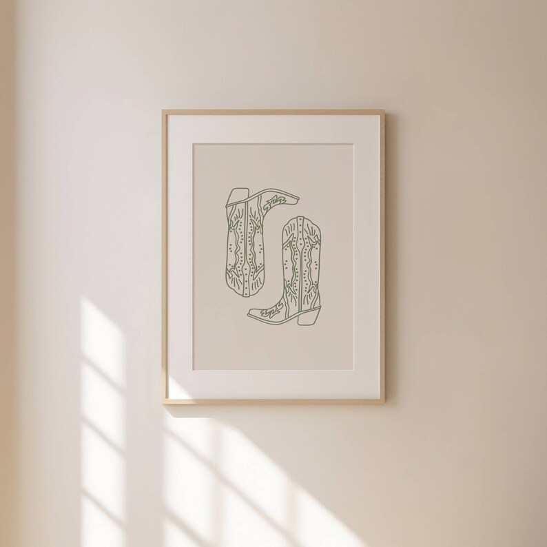 May include: A framed print showing a minimalist illustration of two cowboy boots. The boots are drawn in a muted green hue against a neutral backdrop. The artwork is enclosed in a light-coloured wooden frame, ideal for home decor.