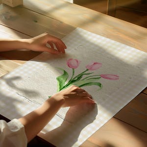 Pode incluir: Uma pe&ccedil;a retangular de papel texturizado branco sujo com uma pintura em aquarela de tr&ecirc;s tulipas cor-de-rosa com caules e folhas verdes. A obra est&aacute; centrada numa superf&iacute;cie aos quadrados bege claro e branco com sombras de luz solar.
