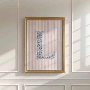 Op de afbeelding: Een ingelijst kunstwerk met de letter "L" in een blauw bloemenpatroon. De achtergrond heeft roze en witte verticale strepen. De gouden lijst voegt een vleugje elegantie toe aan dit decoratieve stuk.