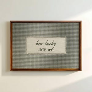 May include: A framed wall art piece with a brown wooden frame. The background features a gray and white gingham pattern. A rectangular patch with the words "how lucky are we" is centered on the artwork.