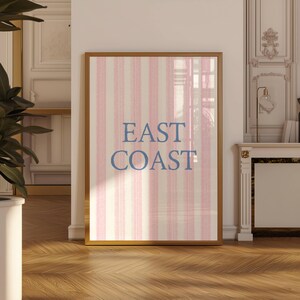 May include: Framed artwork with a gold border. The artwork features a vertical striped pattern in pink and off-white. The words "EAST COAST" are printed in blue, centered on the artwork.