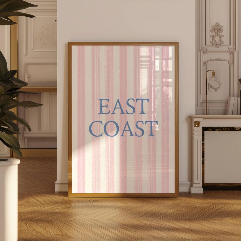 May include: Framed artwork with a gold border. The artwork features a vertical striped pattern in pink and off-white. The words "EAST COAST" are printed in blue, centered on the artwork.