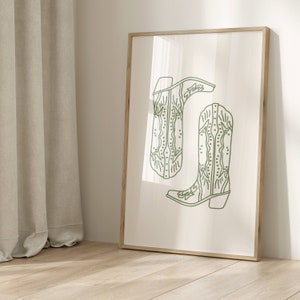 May include: A framed art print with a minimalist design of two cowboy boots in light green. The boots are illustrated with detailed stitching and a classic western style, set against a cream background. The frame is made of wood.