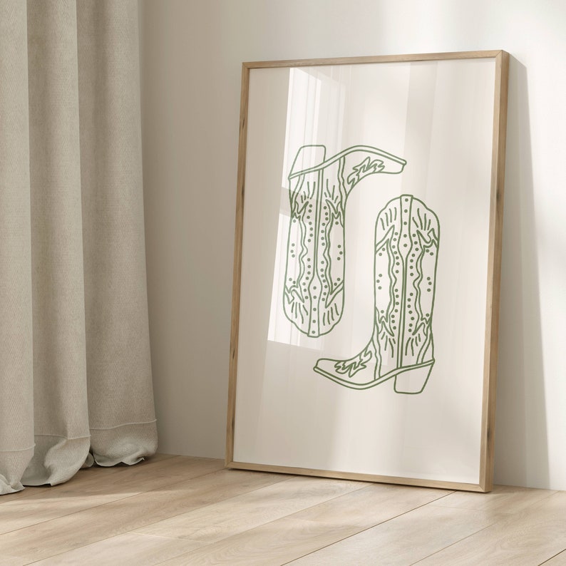 May include: A framed art print with a minimalist design of two cowboy boots in light green. The boots are illustrated with detailed stitching and a classic western style, set against a cream background. The frame is made of wood.