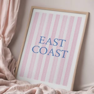 May include: Framed art print with a light wood frame. The artwork features vertical pink and white stripes with the words "EAST COAST" in blue lettering. The print is displayed on a pink fabric background.