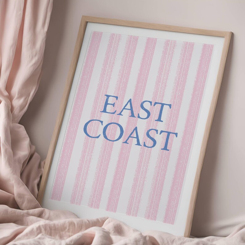 May include: Framed art print with a light wood frame. The artwork features vertical pink and white stripes with the words "EAST COAST" in blue lettering. The print is displayed on a pink fabric background.
