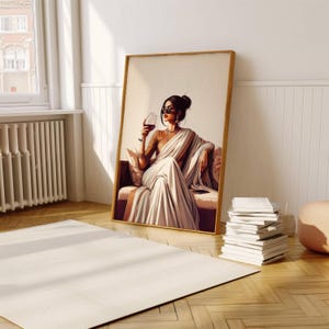 Puede incluir: Una impresi&oacute;n art&iacute;stica enmarcada representa a una mujer con un sari blanco, sentada en un sof&aacute;, sosteniendo una copa de vino tinto. La obra de arte tiene un marco dorado y est&aacute; apoyada contra una pared blanca. La habitaci&oacute;n tiene una alfombra y una pila de libros.