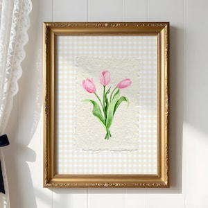 Pode incluir: Uma moldura dourada ornamentada retangular vertical contendo uma pintura em aquarela de tr&ecirc;s tulipas cor-de-rosa com caules e folhas verdes em papel texturizado branco sujo, sobre um fundo com padr&atilde;o xadrez vichy bege claro e branco.
