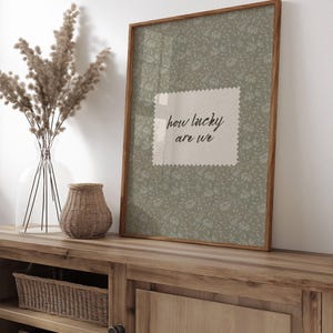 May include: A framed print with the text "how lucky are we" on a light green floral background. The frame is made of wood. A vase with dried plants and a woven basket are on a wooden cabinet.