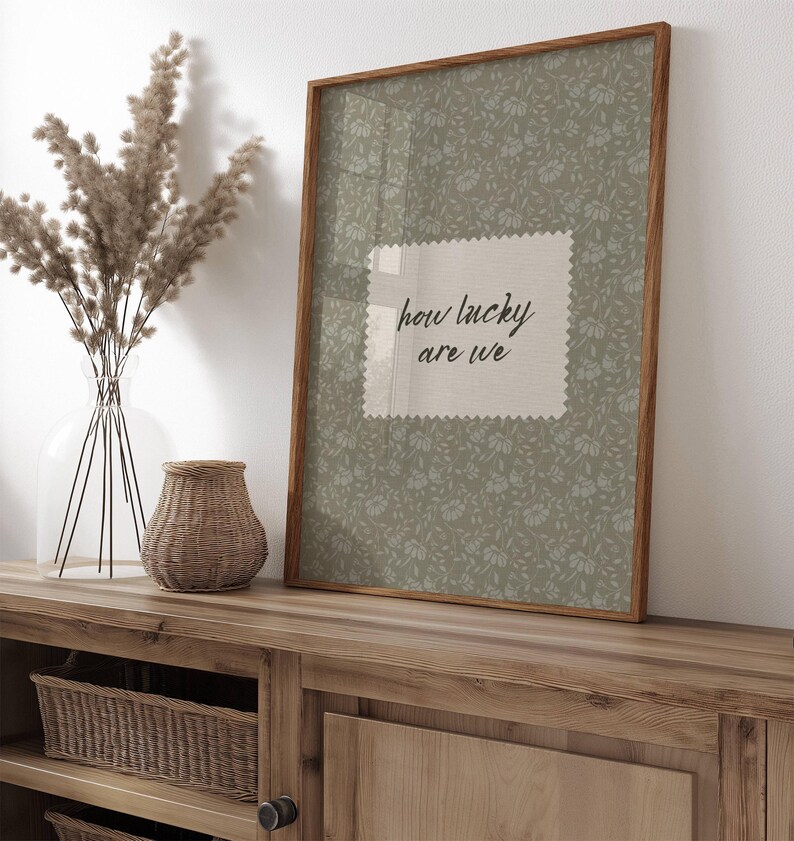 May include: A framed print with the text "how lucky are we" on a light green floral background. The frame is made of wood. A vase with dried plants and a woven basket are on a wooden cabinet.