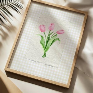 Pode incluir: Uma pe&ccedil;a de arte de parede emoldurada com uma aguarela de tr&ecirc;s t&uacute;lipas cor-de-rosa com caules e folhas verdes em papel texturado com bordas irregulares. A obra est&aacute; sobre um fundo axadrezado bege claro e branco numa moldura de madeira clara.