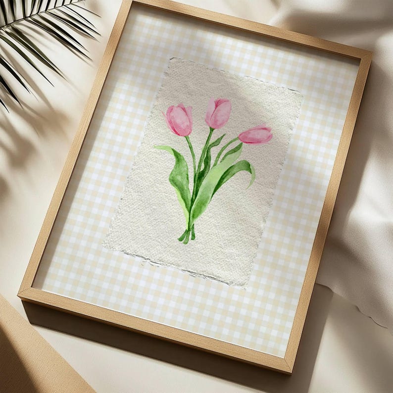 Pode incluir: Uma pe&ccedil;a de arte de parede emoldurada com uma aguarela de tr&ecirc;s t&uacute;lipas cor-de-rosa com caules e folhas verdes em papel texturado com bordas irregulares. A obra est&aacute; sobre um fundo axadrezado bege claro e branco numa moldura de madeira clara.