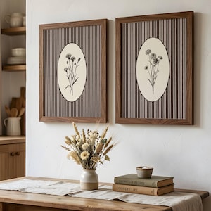 May include: Two rectangular wall art pieces with dark wood frames, featuring a vertical pinstripe background and a central cream-coloured oval containing a black and white botanical illustration of wildflowers, displayed side by side on a wall.