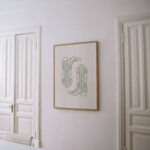 May include: A framed print showing two green cowboy boots in a line drawing style. The boots have decorative stitching and are set against a white background. The artwork is displayed on a white wall, adding a touch of rustic charm.