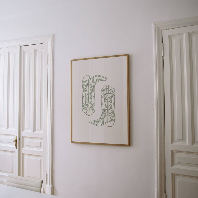 May include: A framed print showing two green cowboy boots in a line drawing style. The boots have decorative stitching and are set against a white background. The artwork is displayed on a white wall, adding a touch of rustic charm.