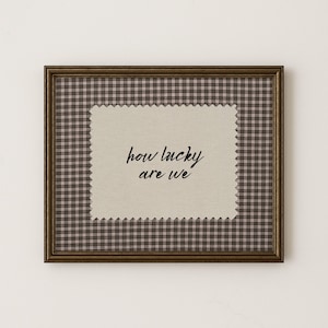 May include: A framed artwork with a brown plaid pattern background and a beige fabric insert. The insert features the phrase "how lucky are we" in black script. The frame is a dark brown color.
