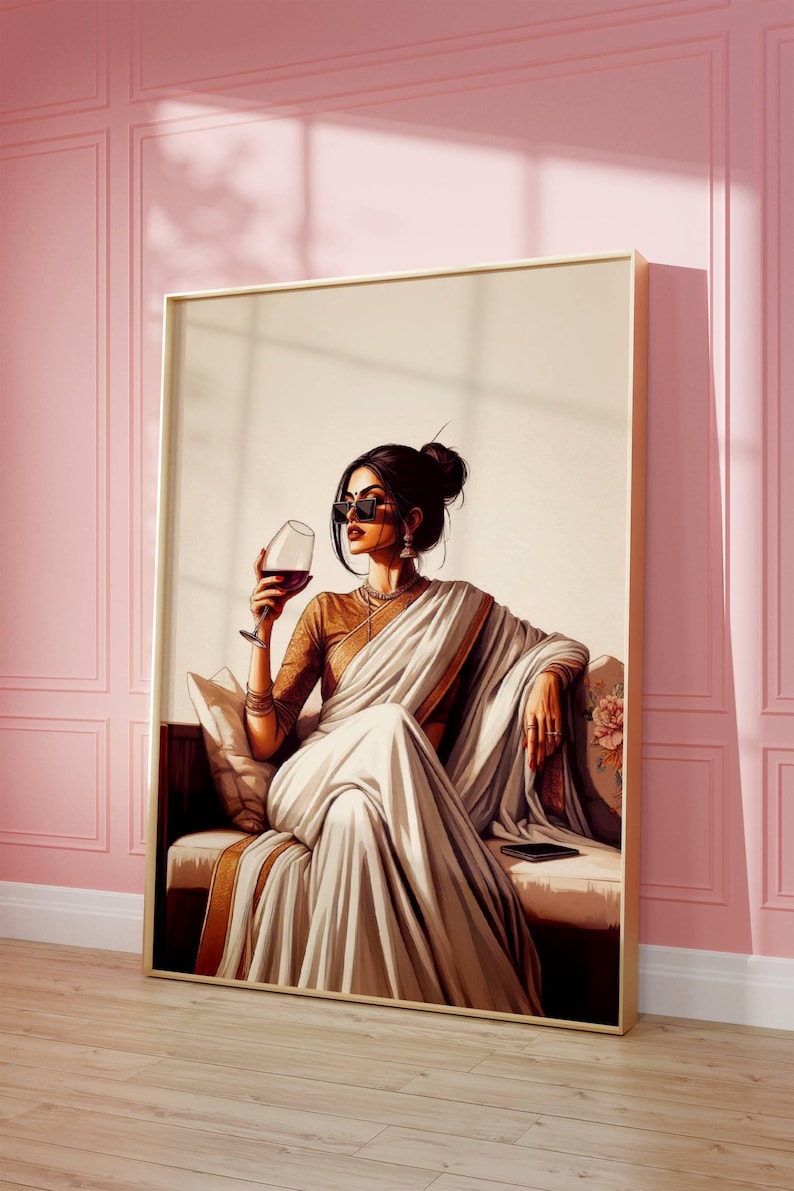 Puede incluir: Una obra de arte enmarcada representa a una mujer con un sari blanco, sosteniendo una copa de vino tinto. Lleva gafas de sol y joyas, sentada en un sof&aacute; con estampado floral. La obra tiene un marco dorado y est&aacute; sobre una pared rosa.