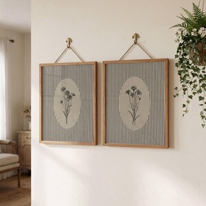 May include: Two square wall art pieces with light wood frames, each featuring a vertical gray and white striped background. Centered on each is an oval beige inset displaying a black line drawing of a wildflower bouquet, hung by a rope.