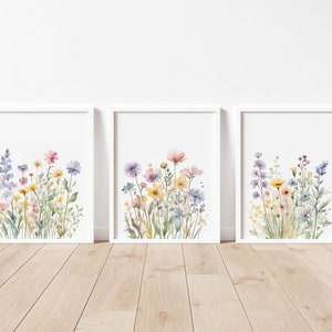 May include: Three framed watercolour prints depicting colourful wildflowers. The artwork showcases a variety of blooms in shades of pink, yellow, blue, and purple, set against a white background. The frames are white, displayed on a wooden floor.