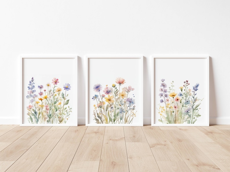 May include: Three framed watercolour prints depicting colourful wildflowers. The artwork showcases a variety of blooms in shades of pink, yellow, blue, and purple, set against a white background. The frames are white, displayed on a wooden floor.