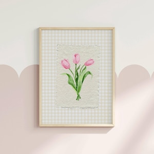 Pode incluir: Uma impress&atilde;o de arte emoldurada com uma aguarela de tr&ecirc;s t&uacute;lipas cor-de-rosa com caules e folhas verdes sobre papel texturado branco sujo. A obra de arte est&aacute; colocada sobre um fundo de xadrez vichy bege claro e branco numa moldura de madeira.