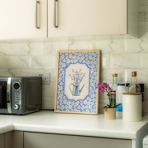 May include: A framed print featuring a blue watering can with lavender, set against a blue floral background. The print is in a light wood frame and sits on a white countertop in a kitchen setting, next to a microwave and other kitchen items.