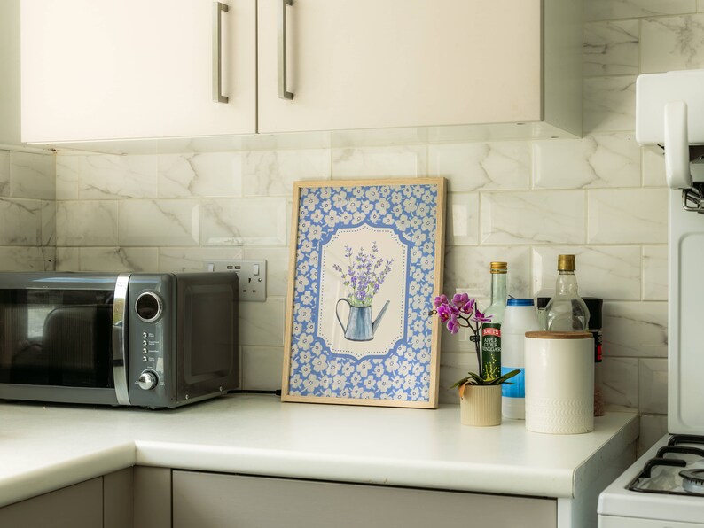 May include: A framed print featuring a blue watering can with lavender, set against a blue floral background. The print is in a light wood frame and sits on a white countertop in a kitchen setting, next to a microwave and other kitchen items.