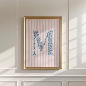 Könnte beinhalten: Gerahmter Kunstdruck mit dem Buchstaben "M" in blauem Blumenmuster. Der Hintergrund hat rosa und weiße vertikale Streifen. Das Kunstwerk befindet sich in einem goldenen Rahmen. Das Thema ist dekorativ und eignet sich für die Inneneinrichtung.