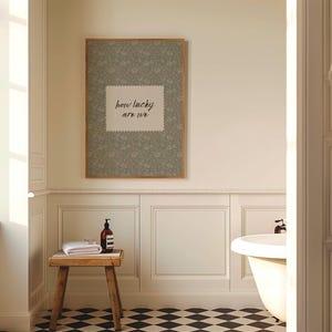 May include: A bathroom scene featuring a framed print with the text "how lucky are we" above a wooden stool. The stool holds a rolled white towel and a brown bottle. The room has a black and white checkered floor.