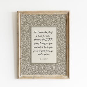 May include: A framed print featuring a floral patterned border and a cream-coloured centre. The text reads "For I know the plans I have for you..." in black script. The frame is a light, distressed wood colour.