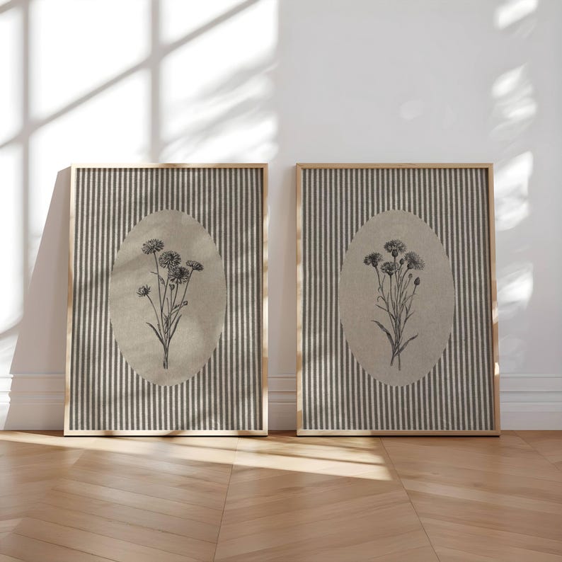 May include: Two rectangular framed wall art pieces featuring a vertical dark gray and light gray striped background. Each frame contains a centered beige oval with a detailed black ink botanical illustration of a wildflower bouquet inside.