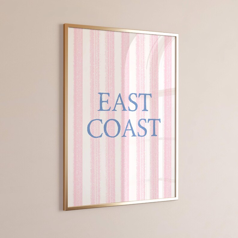 May include: Framed wall art with a gold-colored frame. The artwork features vertical stripes in pink and white, with the words "EAST COAST" in blue, centered on the design. The art is displayed on a light pink wall.