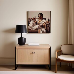 May include: A framed artwork depicts a person in a white sari, seated on a brown sofa, holding a drink. A black lamp sits on a light wood cabinet with black legs. A white book is on the cabinet. A wooden chair is to the right.