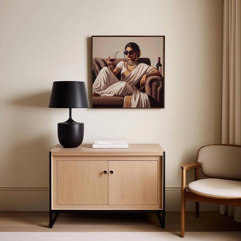 May include: A framed artwork depicts a person in a white sari, seated on a brown sofa, holding a drink. A black lamp sits on a light wood cabinet with black legs. A white book is on the cabinet. A wooden chair is to the right.