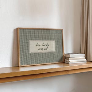 May include: A framed artwork with a wooden frame. The artwork features a gingham pattern in shades of gray and white. A rectangular patch with the words "how lucky are we" is centered on the artwork. A stack of books sits to the right.