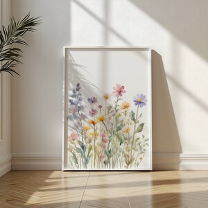 May include: A framed watercolour print showcasing a colourful selection of wildflowers in pink, yellow, purple, and blue hues. The artwork is set against a white backdrop, within a white frame. The print is leaning against a white wall.