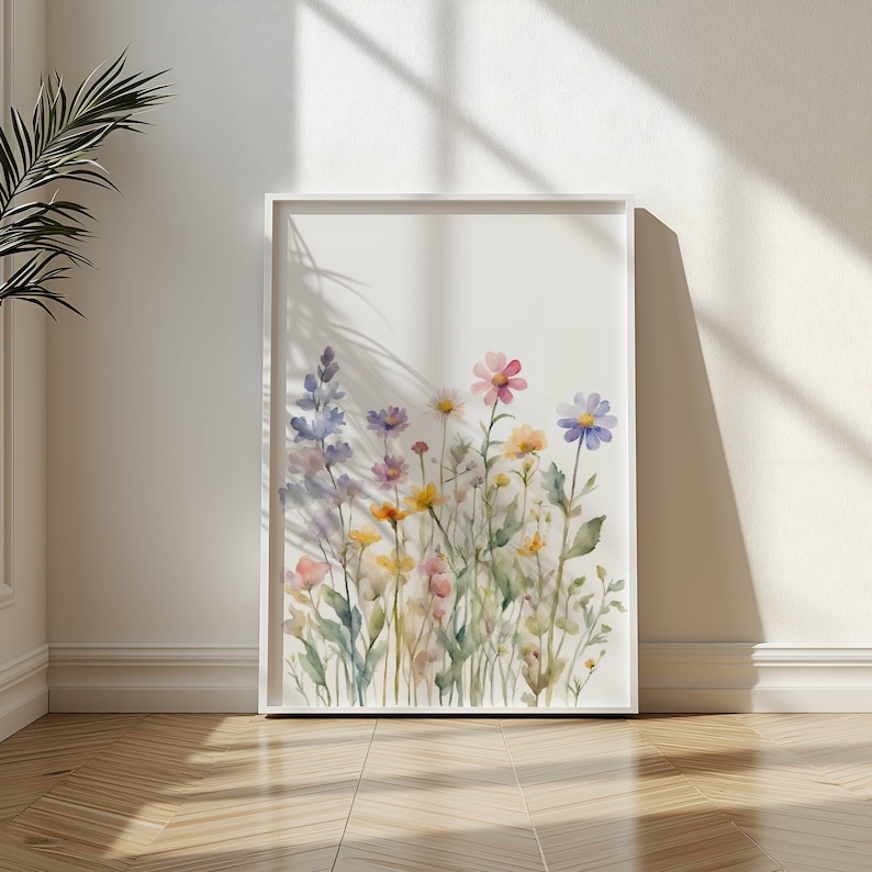 May include: A framed watercolour print showcasing a colourful selection of wildflowers in pink, yellow, purple, and blue hues. The artwork is set against a white backdrop, within a white frame. The print is leaning against a white wall.