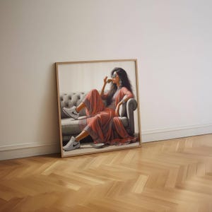 May include: A framed artwork depicts a person in a coral-colored outfit, seated on a gray tufted sofa, sipping from a cup. The person wears sunglasses and white sneakers. The artwork is leaning against a white wall on a wooden floor.