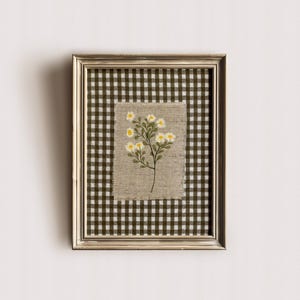 May include: A framed floral embroidery. The artwork features a sprig of yellow and white flowers on a beige linen background. The linen is set against a green and white checkered fabric, all within a silver-coloured frame.